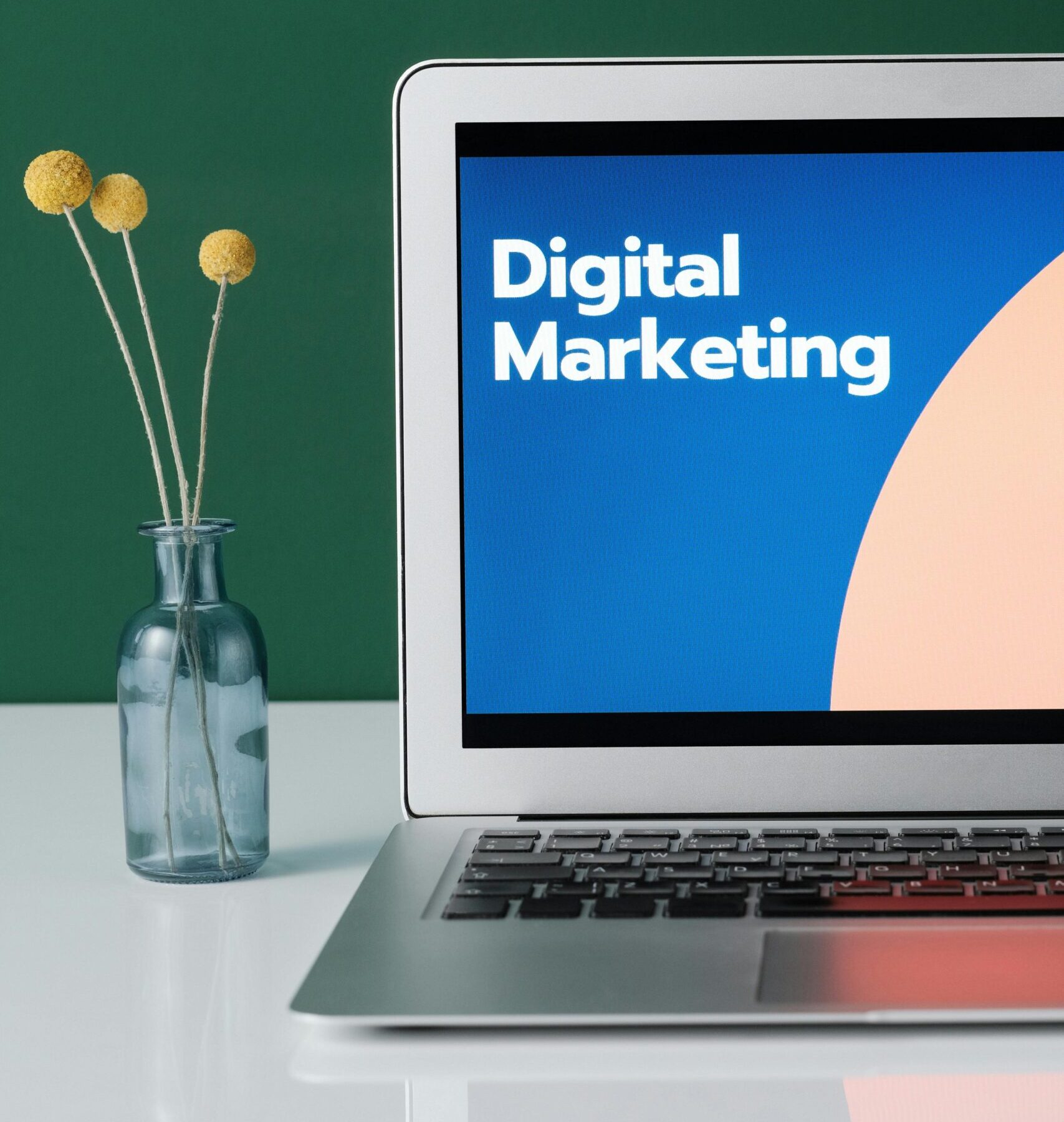 A laptop displaying 'Digital Marketing' with a minimalist floral decor in a glass vase.